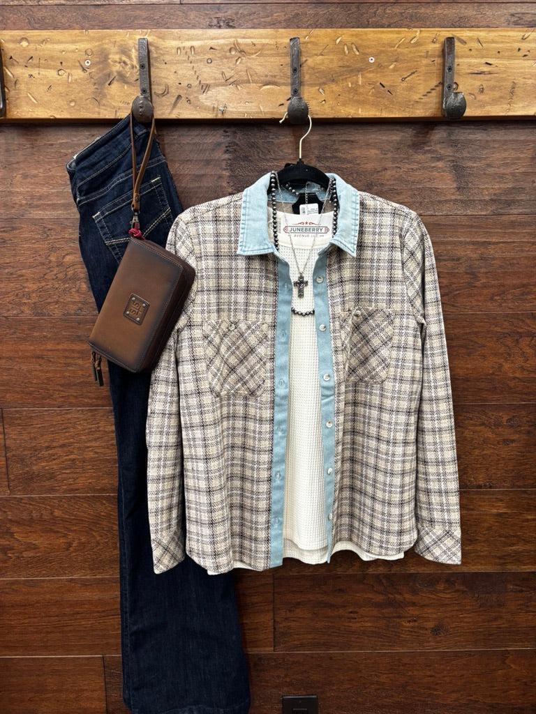 A plaid button-up shirt with a light blue denim collar and cuffs layered over a white knit shirt, hanging on a wooden wall rack next to dark blue jeans and a brown leather pouch.