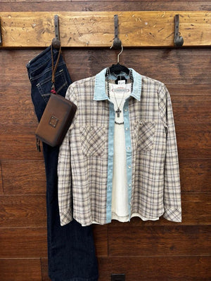 A plaid button-up shirt with a light blue denim collar and cuffs layered over a white knit shirt, hanging on a wooden wall rack next to dark blue jeans and a brown leather pouch.