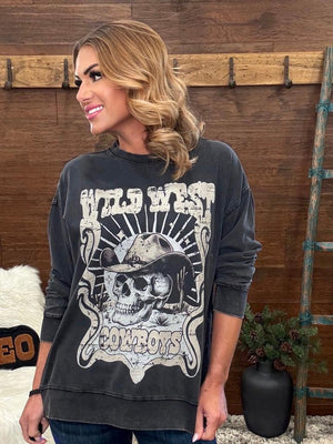 A woman wearing a dark gray washed graphic sweatshirt with a skull and cowboy hat design that reads 'Wild West Cowboys', posed in front of a rustic wooden wall.