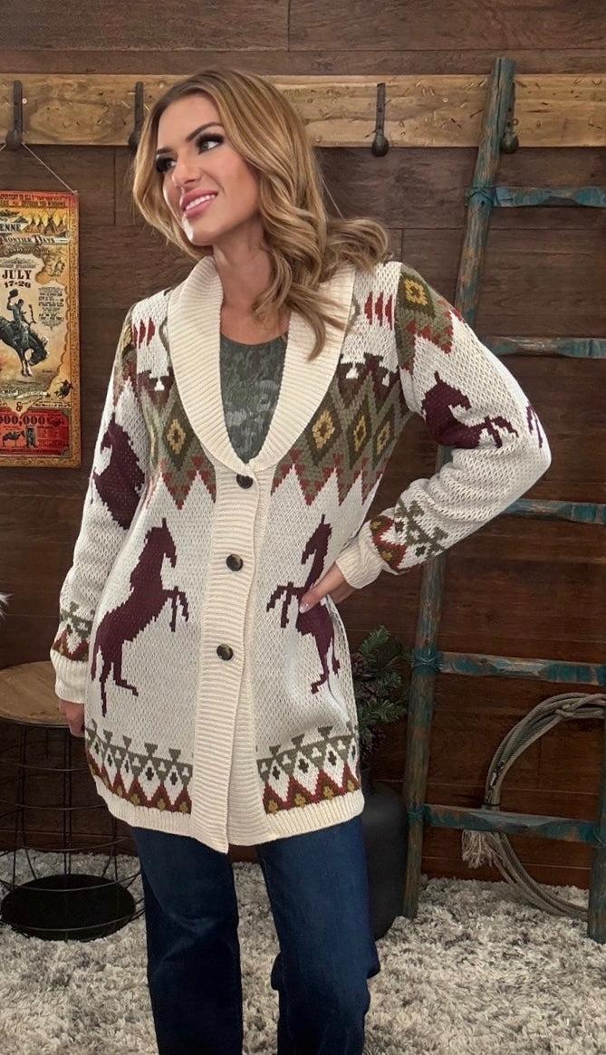 A woman models a cream knit cardigan with brown horse motifs and geometric patterns, paired with dark blue jeans in a rustic indoor setting with a wooden wall and ladder.