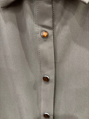 A taupe long-sleeve button-down blouse hanging on a black hanger against a rustic wooden wall, styled with dark blue jeans and a cream crossbody bag on hooks.