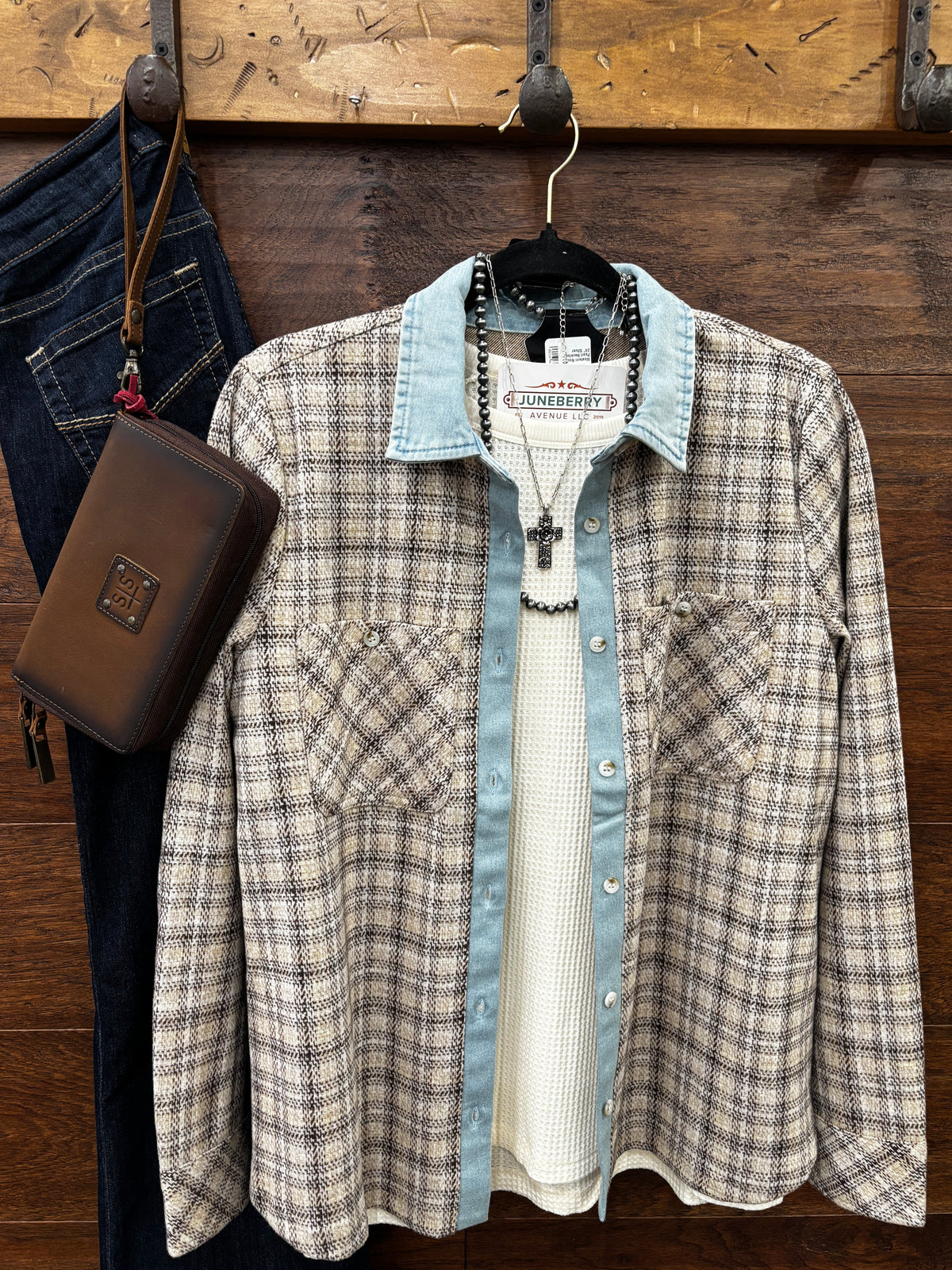 A plaid button-up shirt with a light blue denim collar and cuffs layered over a white knit shirt, hanging on a wooden wall rack next to dark blue jeans and a brown leather pouch.