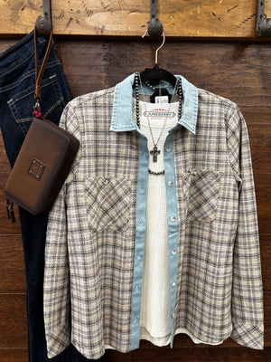 A plaid button-up shirt with a light blue denim collar and cuffs layered over a white knit shirt, hanging on a wooden wall rack next to dark blue jeans and a brown leather pouch.