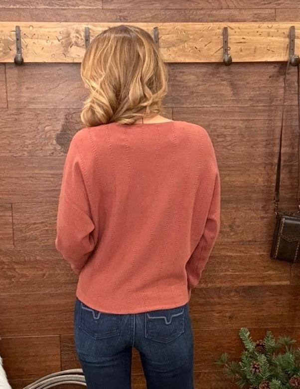 A woman viewed from behind wearing a dusty rose long-sleeve knit sweater and blue jeans, standing in front of a rustic wooden wall with pegs.