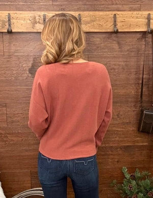 A woman viewed from behind wearing a dusty rose long-sleeve knit sweater and blue jeans, standing in front of a rustic wooden wall with pegs.