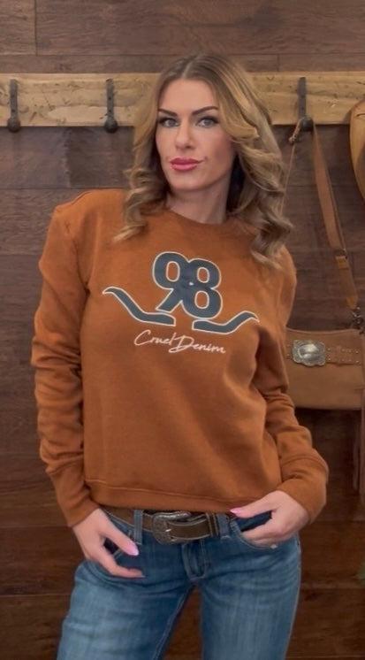 Woman wearing a brown sweatshirt with a logo, standing against a wooden wall.