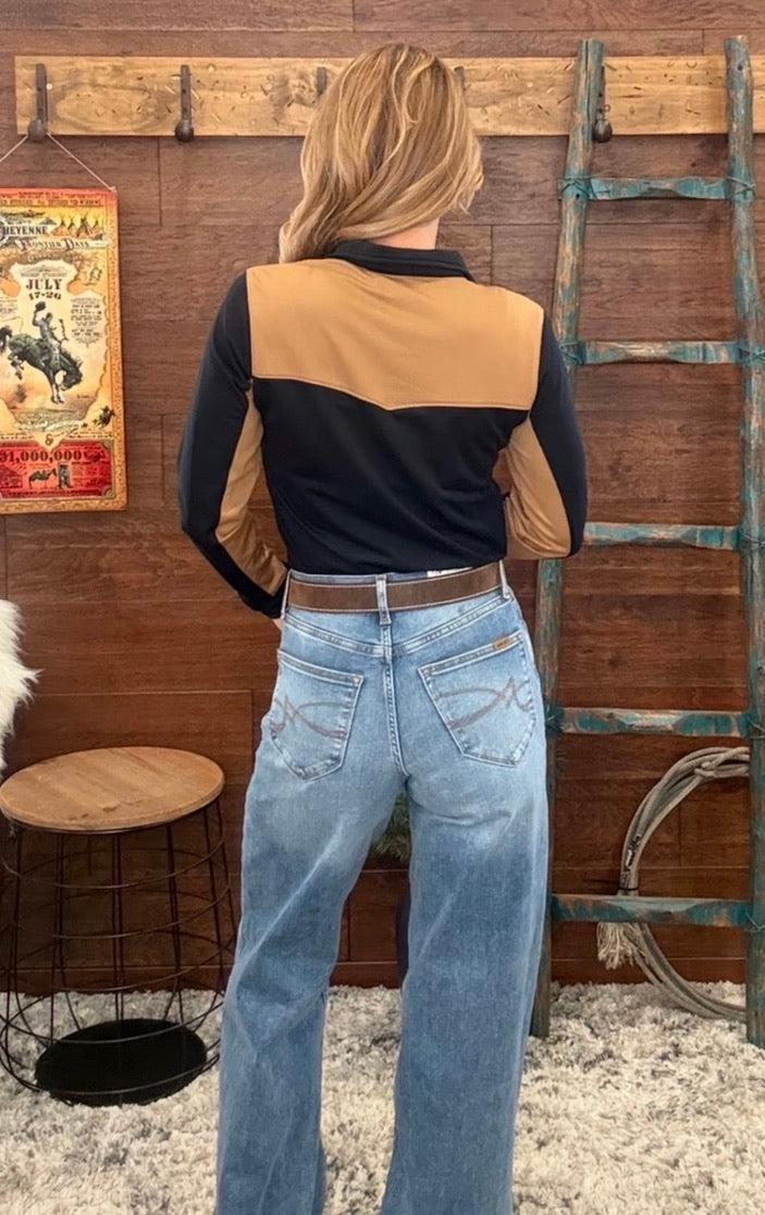 A woman viewed from behind wearing a color-block long-sleeve shirt with tan and black panels, paired with light-wash blue jeans, standing in a rustic room with a wooden wall, ladder, and cowboy poster.