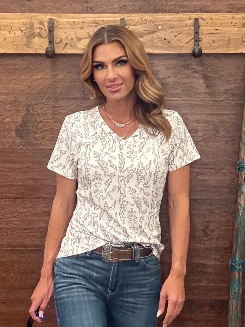 Woman wearing a white patterned shirt and blue jeans standing against a wooden wall.