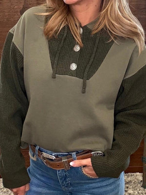A woman wearing a color-blocked henley sweater with an olive green body and dark olive green ribbed sleeves and yoke, paired with blue jeans and a brown leather belt.