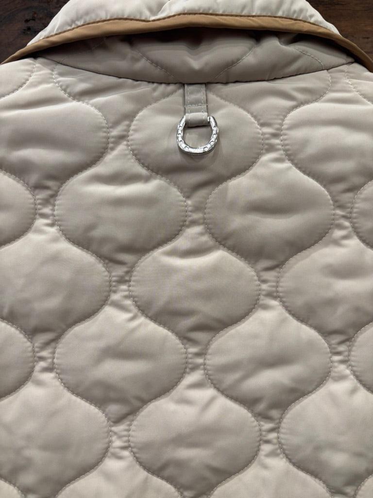 A quilted sleeveless vest in off-white with tan trim, layered over an olive green button-up shirt, hanging on a gray ARIAT branded hanger against a dark wood background.