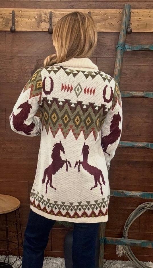 A woman viewed from behind wearing a cream knit cardigan with Southwestern-style geometric patterns and dark red horse motifs, standing in front of a rustic wooden wall with a ladder.