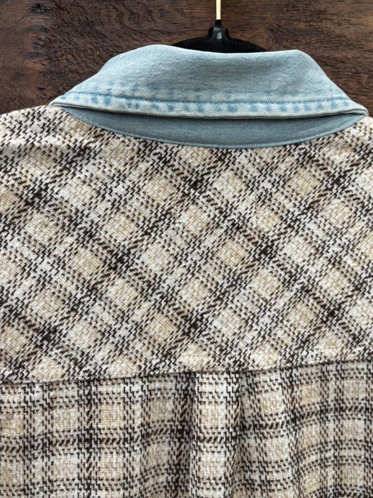A close-up of a plaid tweed jacket or shirt jacket with a light blue denim collar, displayed on a hanger against a dark wood background.