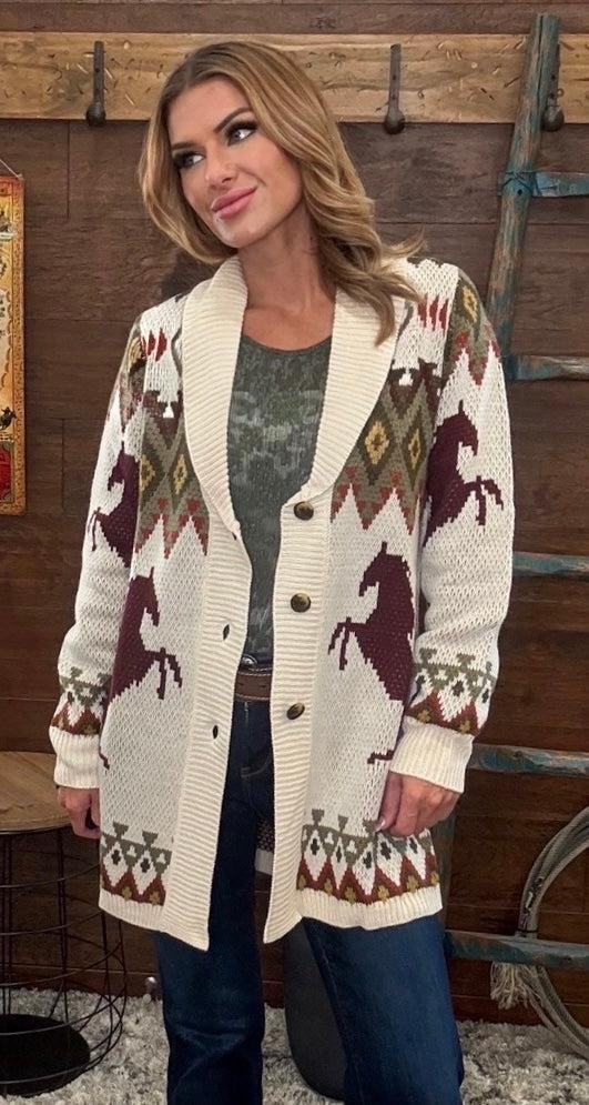 A woman wearing a cream knit cardigan with brown and burgundy horse and geometric patterns, styled over a gray top and blue jeans, standing in front of a rustic wooden wall.