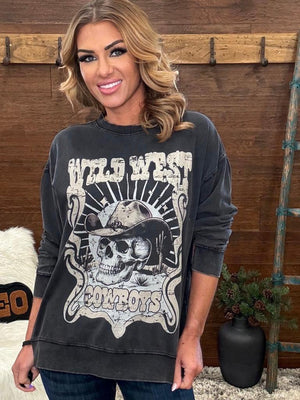 A woman wearing a faded black long-sleeve graphic sweatshirt with a distressed white 'Wild West Cowboys' skull and cowboy hat design, posed in front of a rustic wooden wall.
