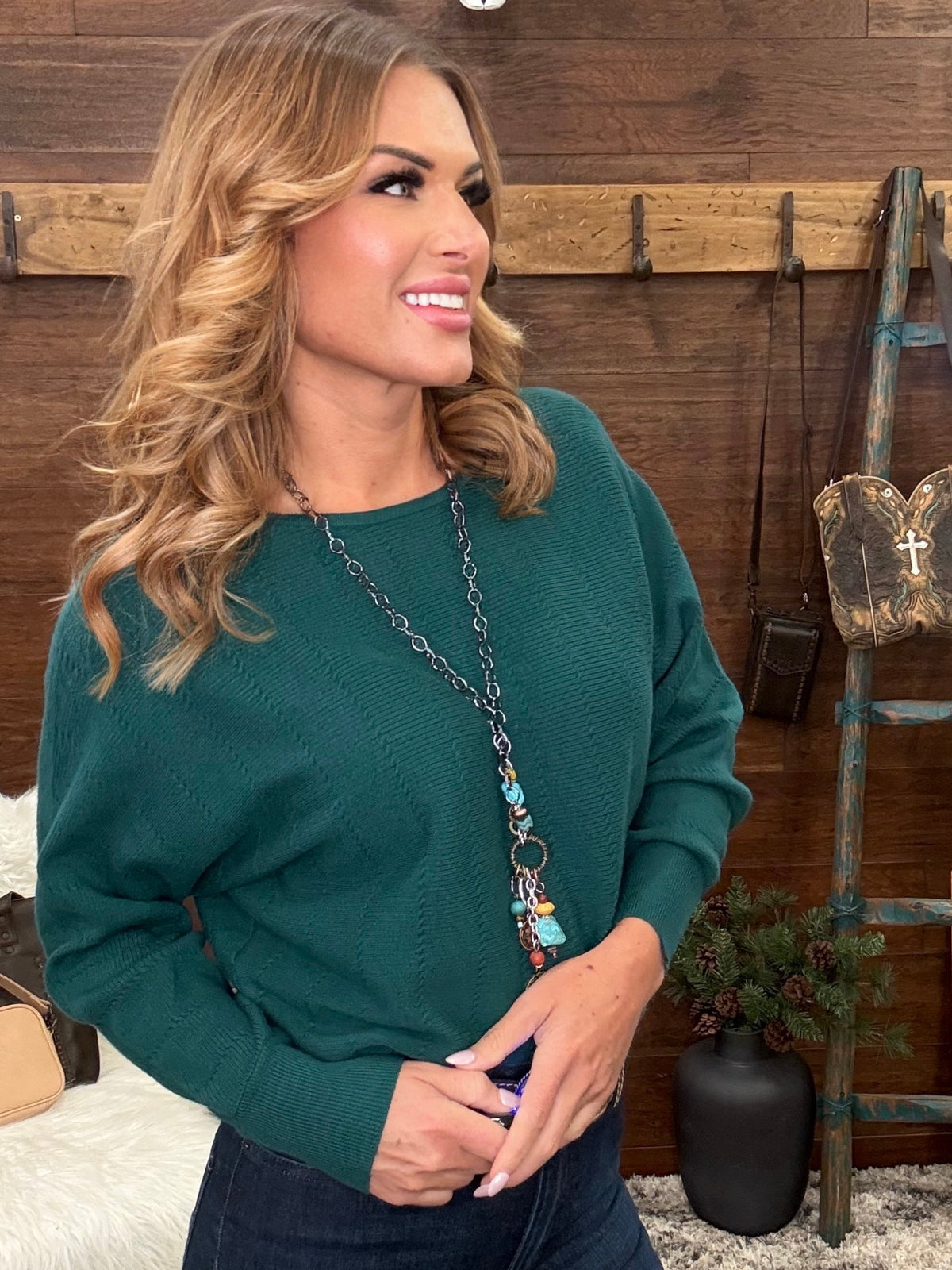 A woman with blonde wavy hair wearing a dark green long-sleeve knit sweater and a long silver chain necklace with colorful beads, standing in front of a rustic wooden wall.