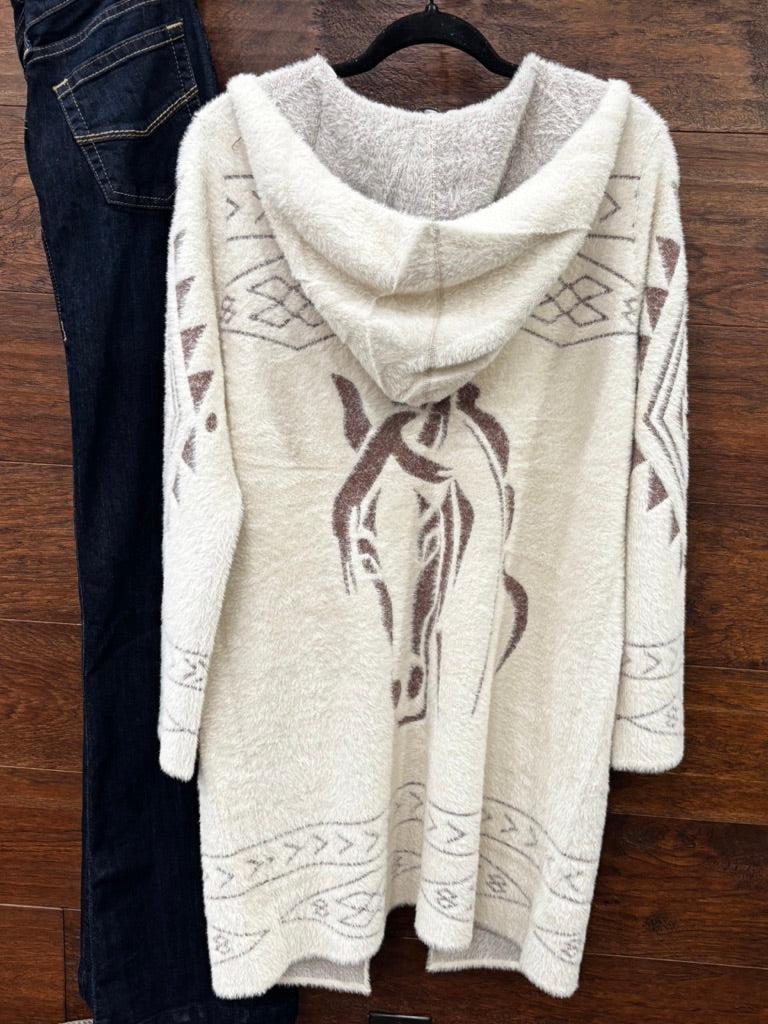 A cream-colored fuzzy hooded cardigan sweater with brown horse and geometric patterns, hanging on a black hanger next to a pair of dark blue jeans against a wooden background.