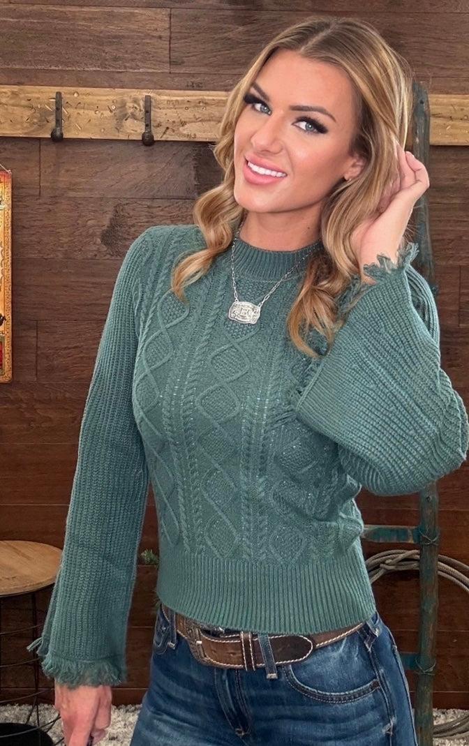 A woman wearing a sage green cable-knit sweater with ruffled cuffs, paired with blue jeans, standing in front of a rustic wooden wall with a ladder.