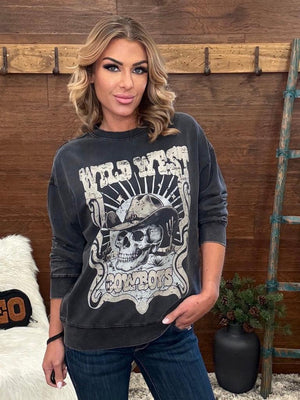 A woman wearing a faded black graphic sweatshirt with a western skull design and the words 'Wild West Cowboys', paired with dark blue jeans, standing in front of a rustic wooden wall.
