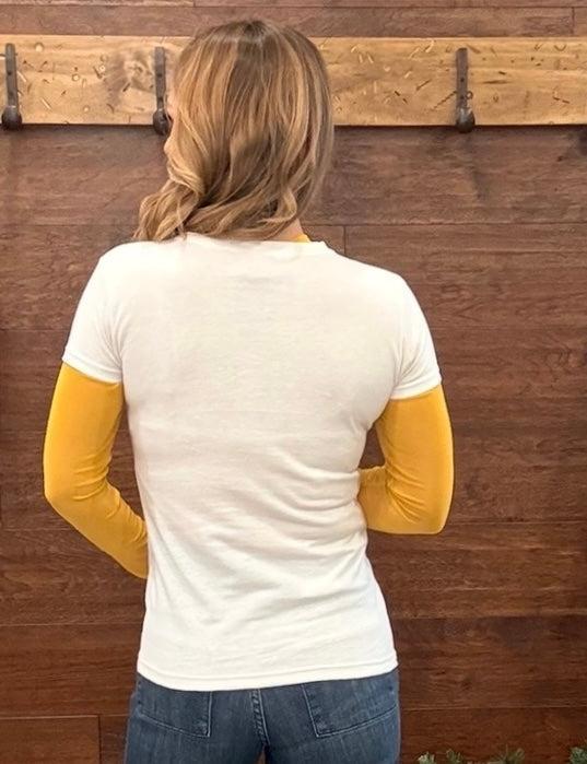 A woman viewed from behind wearing a white short-sleeve t-shirt layered with mustard yellow long sleeves, paired with blue jeans, standing in front of a dark wood wall.