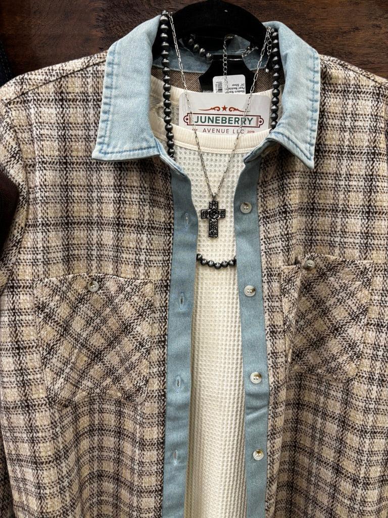 A plaid button-up shirt with a light blue denim collar and cuffs layered over a white knit shirt, hanging on a wooden wall rack next to dark blue jeans and a brown leather pouch.