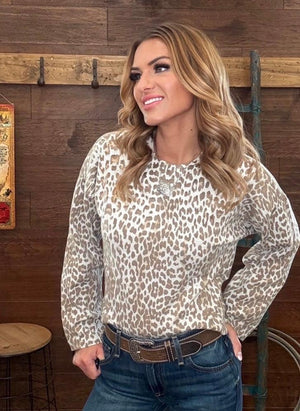 A woman wearing a beige and brown leopard print long-sleeve top with blue jeans, standing in front of a rustic wooden wall.