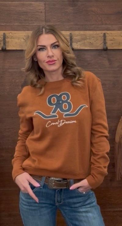Woman wearing a brown sweatshirt with a logo, standing against a wooden wall.