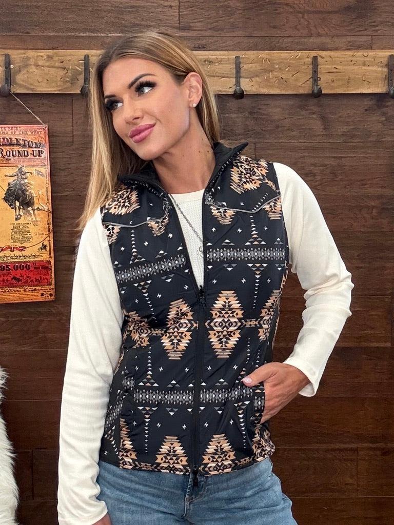 A woman wearing a sleeveless black vest with a beige and white southwestern geometric pattern, layered over a white long-sleeve shirt and blue jeans. 