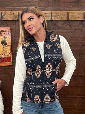 A woman wearing a sleeveless black vest with a beige and white southwestern geometric pattern, layered over a white long-sleeve shirt and blue jeans. 