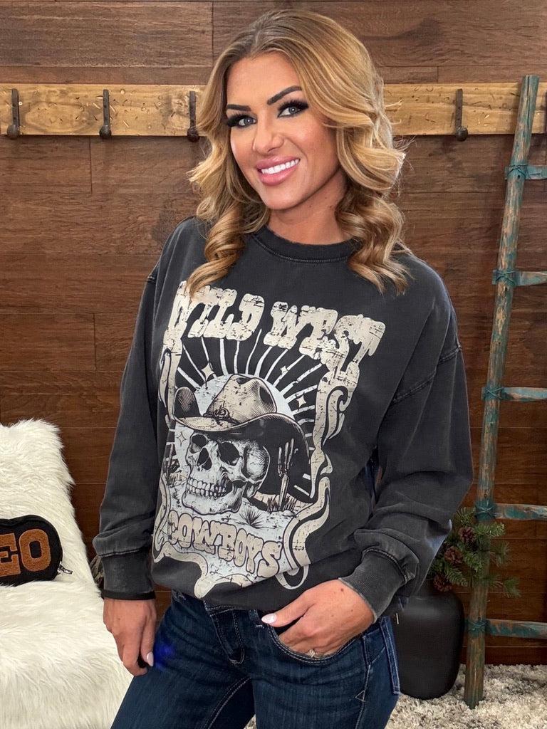 A woman wearing a dark gray washed graphic sweatshirt with a skull and cowboy hat design, paired with dark blue jeans, standing in front of a rustic wooden wall.