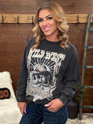 A woman wearing a dark gray washed graphic sweatshirt with a skull and cowboy hat design, paired with dark blue jeans, standing in front of a rustic wooden wall.