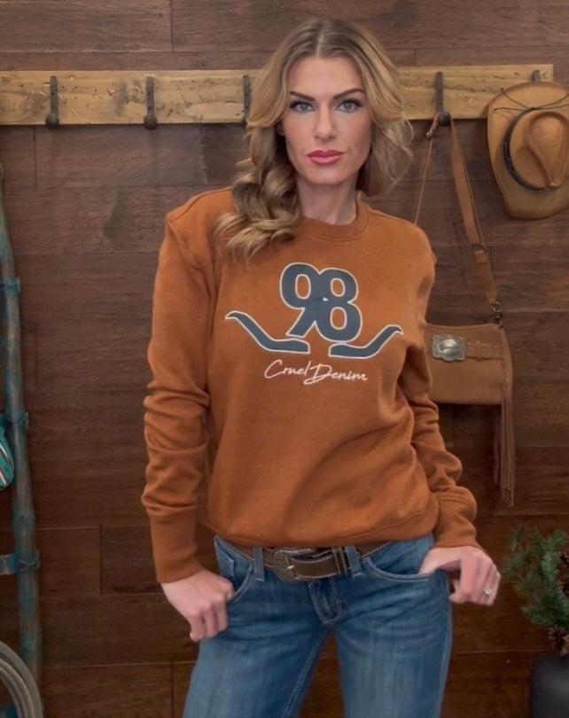 Woman wearing a brown sweatshirt with a logo and text, standing against a wooden wall.