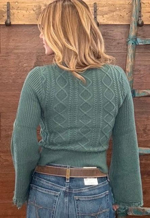 A woman viewed from behind wearing a sage green cable-knit sweater and blue jeans, standing in front of a rustic wooden wall.