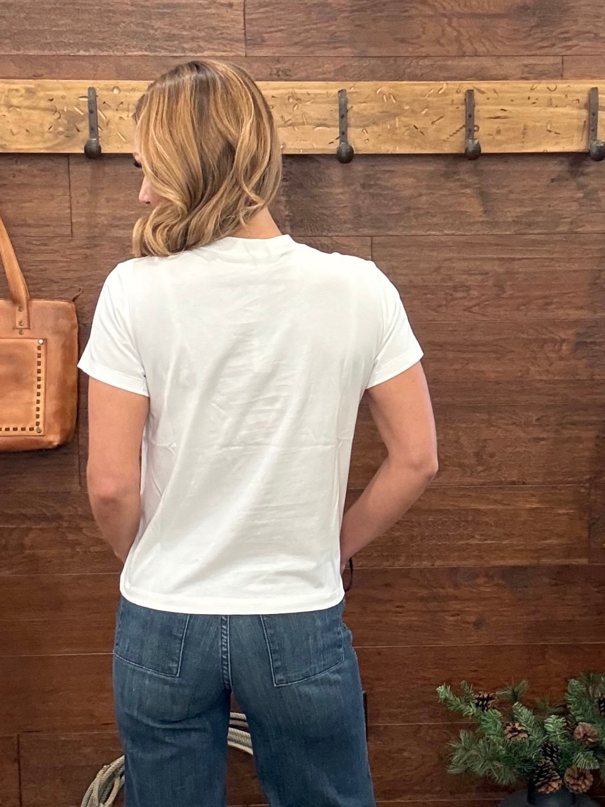 A woman viewed from behind wearing a plain white short-sleeve t-shirt and blue jeans, standing in front of a rustic dark wood wall with a wooden coat rack.