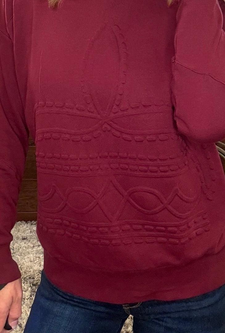 A close-up of a person wearing a burgundy long-sleeve sweater with raised decorative knit patterns across the chest and waist, paired with blue jeans.