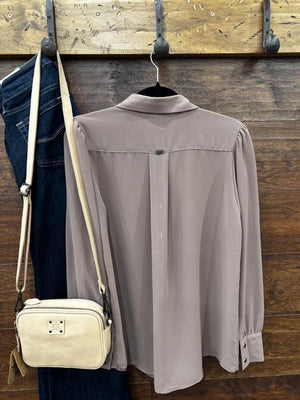 A taupe long-sleeve button-down blouse hanging on a black hanger against a rustic wooden wall, styled with dark blue jeans and a cream crossbody bag on hooks.