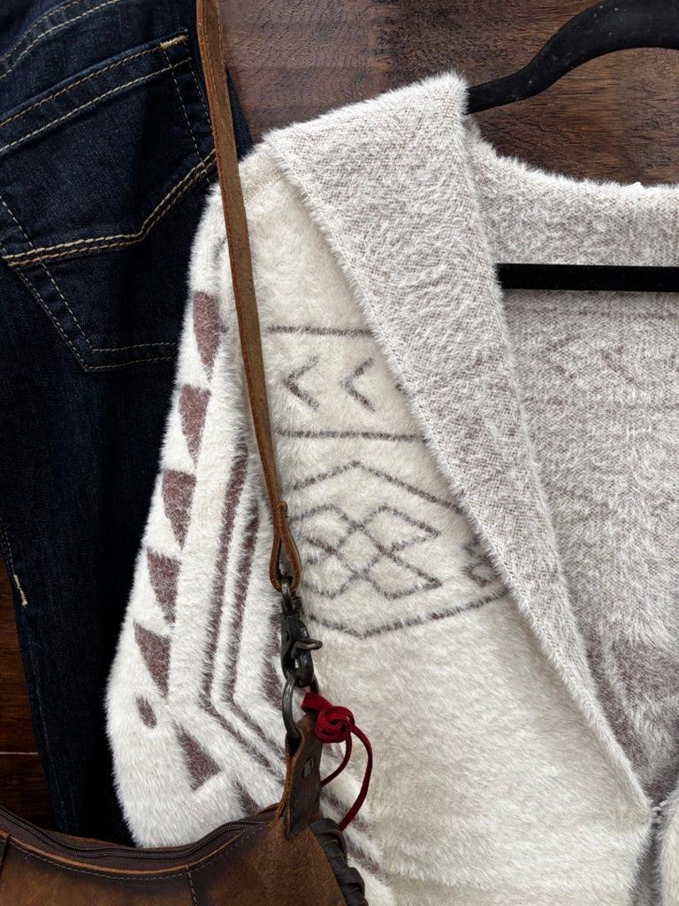 A close-up of a fuzzy knit cardigan with a cream and light gray Southwestern geometric pattern, styled on a hanger next to dark blue jeans and a brown leather shoulder bag.