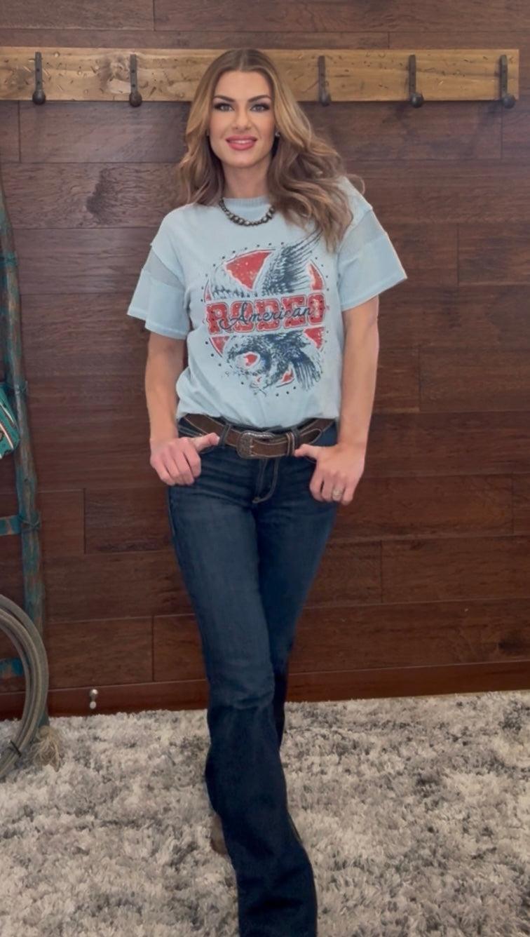Woman wearing a graphic t-shirt and jeans standing against a wooden wall.