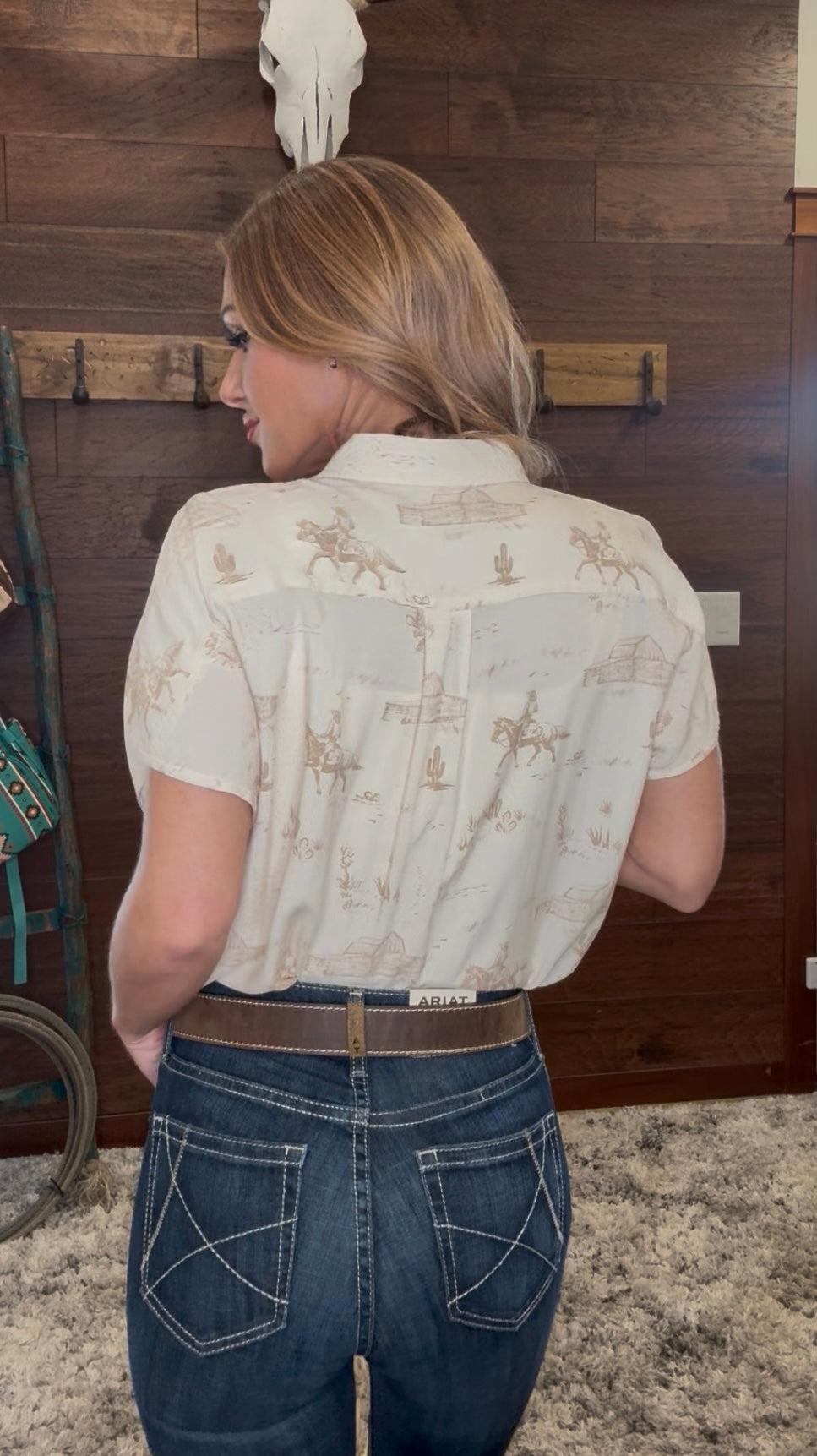 Woman wearing a cream-colored blouse with horse patterns and blue jeans against a wooden wall.