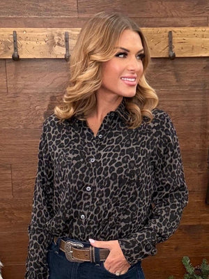 A woman wearing a black and gray leopard print button-down shirt, styled with dark blue jeans and a brown leather belt with a silver buckle, standing in front of a rustic wooden wall.