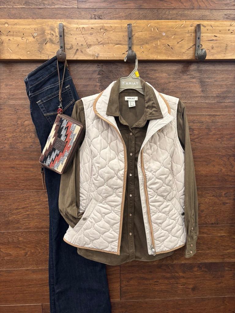 A quilted sleeveless vest in off-white with tan trim, layered over an olive green button-up shirt, hanging on a gray ARIAT branded hanger against a dark wood background.