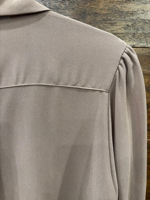 A taupe long-sleeve button-down blouse hanging on a black hanger against a rustic wooden wall, styled with dark blue jeans and a cream crossbody bag on hooks.