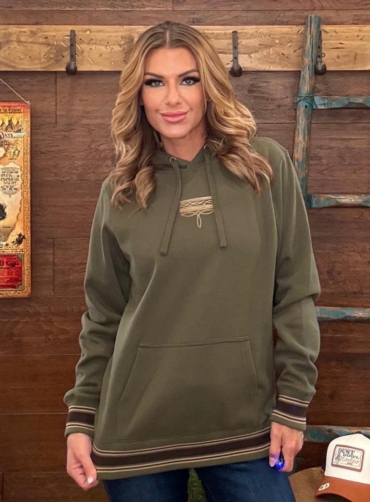 A woman wearing an olive green pullover hoodie with a lace-up chest detail and striped cuffs, standing in front of a rustic wooden wall.