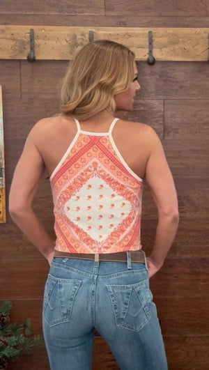 Woman wearing a patterned tank top and blue jeans against a wooden wall.