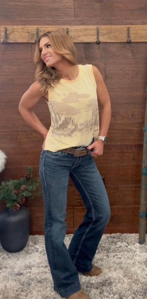 Woman wearing a yellow sleeveless top and blue jeans standing against a wooden wall.
