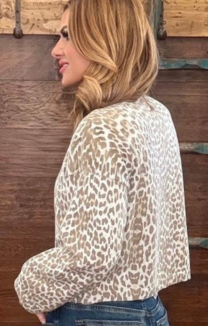 A woman wearing a beige and brown leopard-print long-sleeve top, shown from a side-back angle against a rustic wooden wall.