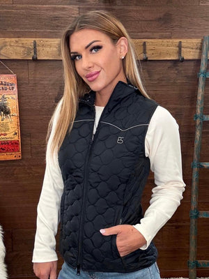 A woman modeling a black quilted sleeveless puffer vest with a small silver logo on the chest, worn over a white long-sleeve shirt and blue jeans. 