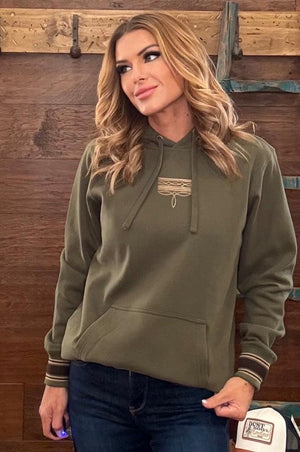 A woman wearing an olive green pullover hoodie with a small embroidered chest graphic and striped cuffs, standing against a rustic wooden wall.