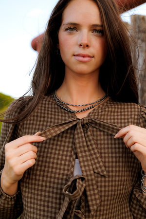 This charming blouse features a timeless brown checkered pattern and a stylish front tie detail, making it a versatile and fashionable addition to any wardrobe.