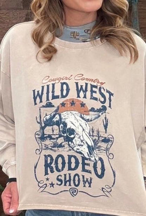 A close-up of a person wearing a cream-colored long-sleeve graphic sweatshirt with a distressed blue and orange 'Wild West Rodeo Show' print on the front.