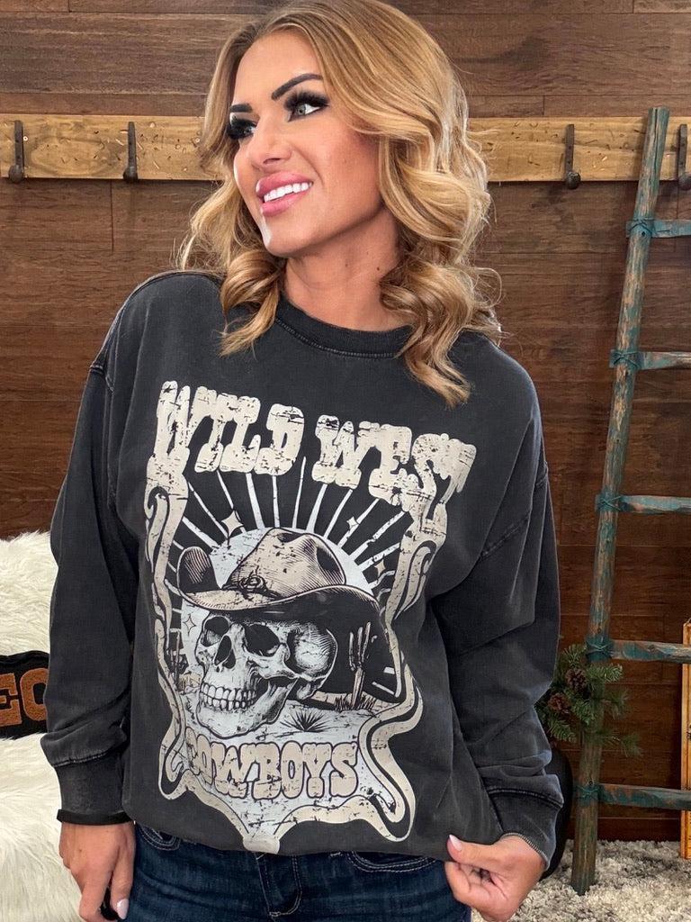 A woman wearing a faded black graphic sweatshirt with a distressed 'Wild West Cowboys' skull design, posed against a rustic wooden wall background.